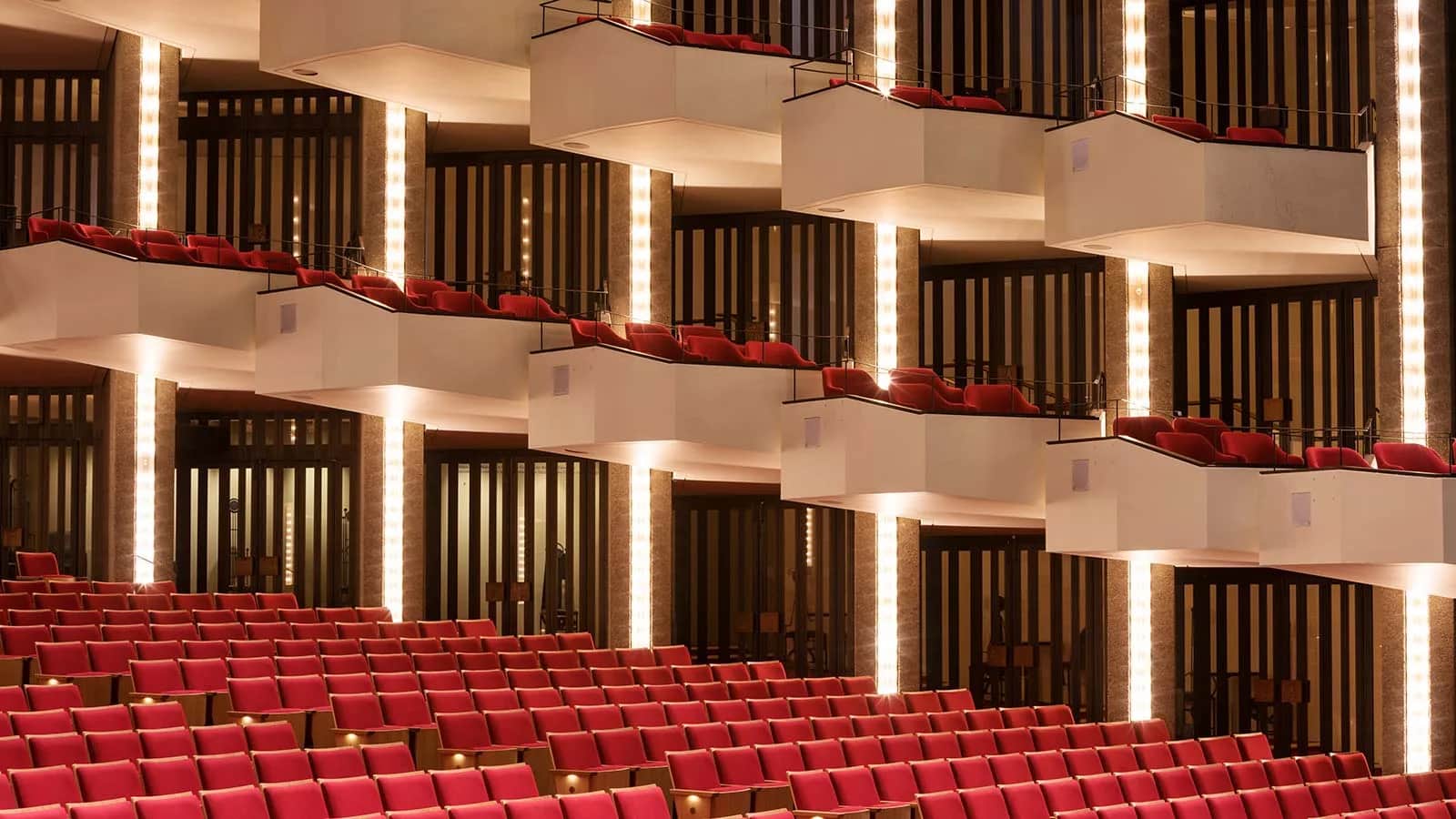 National Arts Centre (NAC), Ottawa: Staggered white balcony fronts with red seats and thin dark guardrails, lit by vertical light strips, above rows of red auditorium seats