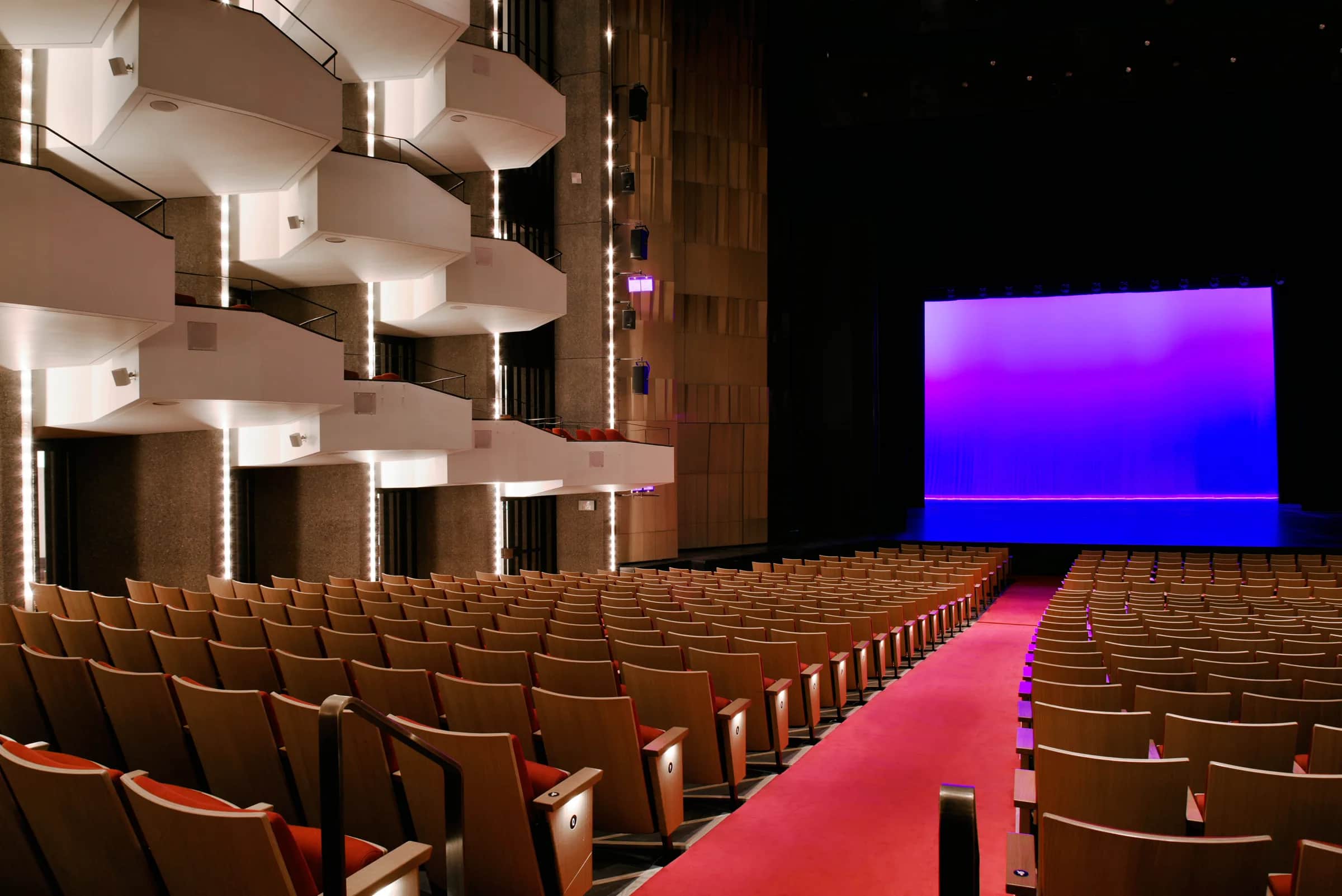 National Arts Centre (NAC), Ottawa: Side balconies with guardrails along their fronts beside rows of seats, a red aisle leading to a stage lit in purple and blue, a dark metal handrail in the foreground