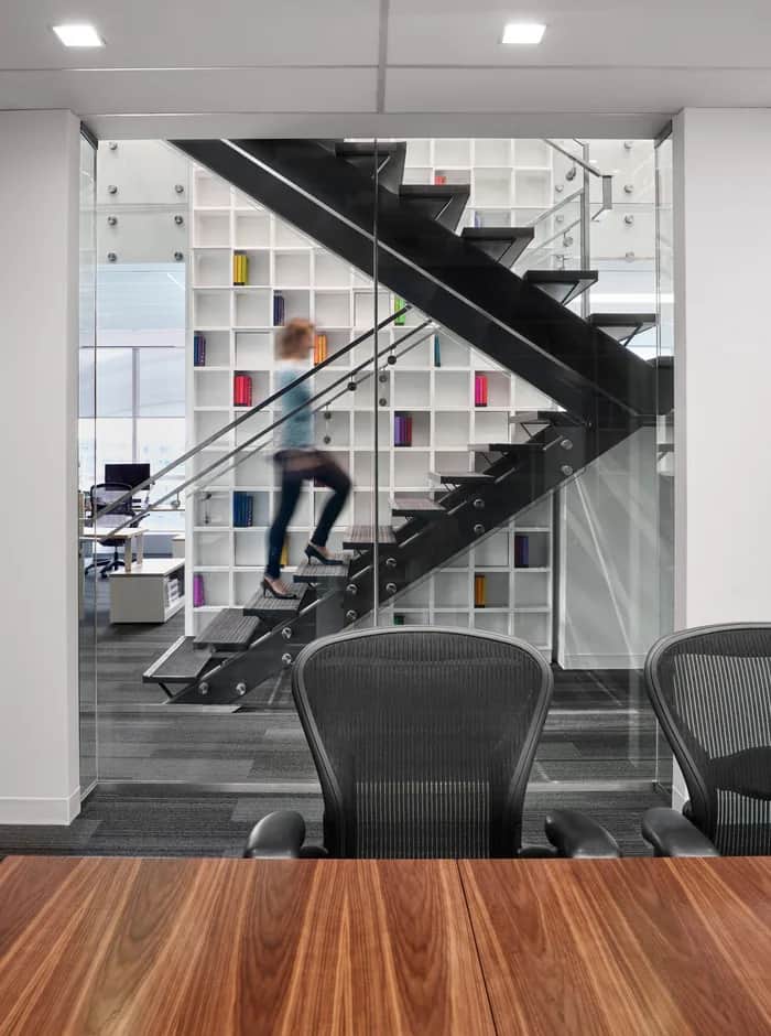 Penguin Stair: Black steel stair with open treads in front of white shelving, seen through a glass meeting-room wall