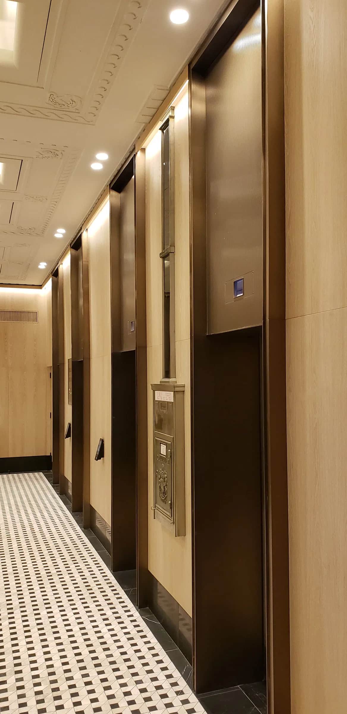 Richmond Elevator Lobby: Row of elevator doors along a lobby wall, with dark bronze surrounds and fluted panels between pale wood-grain walls, a decorative plaster ceiling and a patterned tile floor