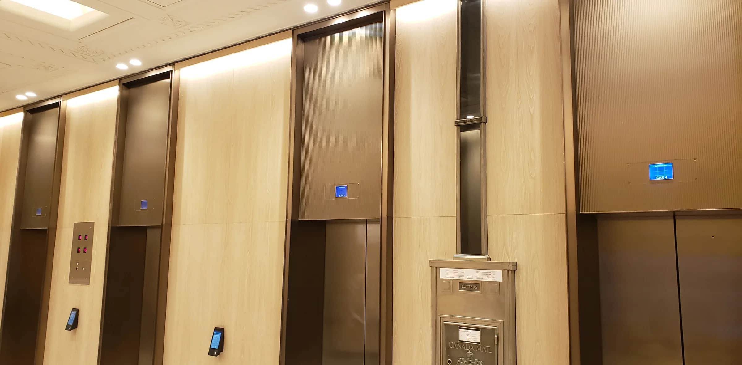 Richmond Elevator Lobby: Elevator lobby wall seen at an angle: fluted bronze panels above the doors and dark bronze surrounds between pale wood-grain panels, with a silver Canada Mail letter box set into the wall