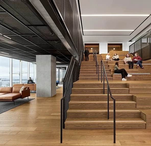 The Well, Toronto: Timber stadium stair with black steel handrails and a mesh guardrail, people seated on the upper steps, with a lounge area and windows beyond