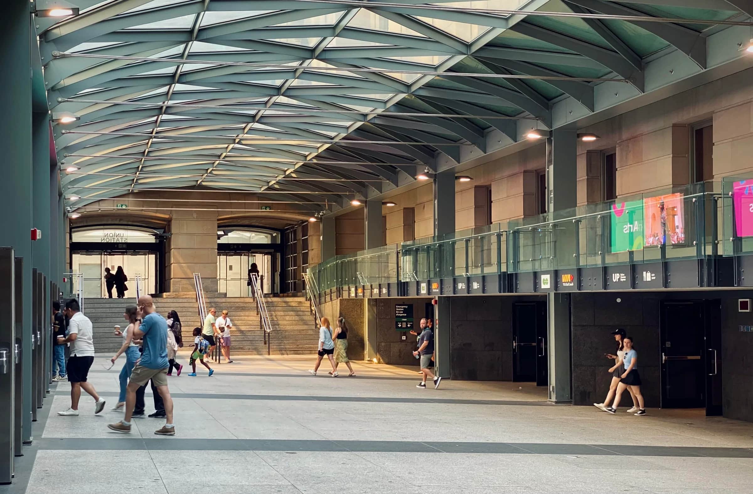 Union Station, Toronto: Stainless steel handrails on a stone stair at the far end of a covered concourse under a glass-and-steel roof, with people walking across the floor