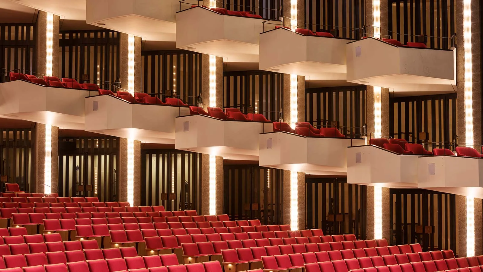 National Arts Centre (NAC), Ottawa: Staggered white balcony fronts with red seats and thin dark guardrails, lit by vertical light strips, above rows of red auditorium seats