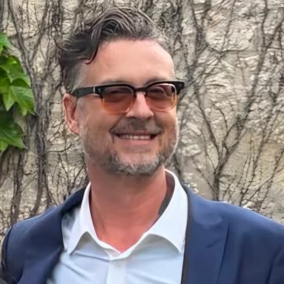 John Day, smiling, in tinted glasses, a white shirt and a navy jacket