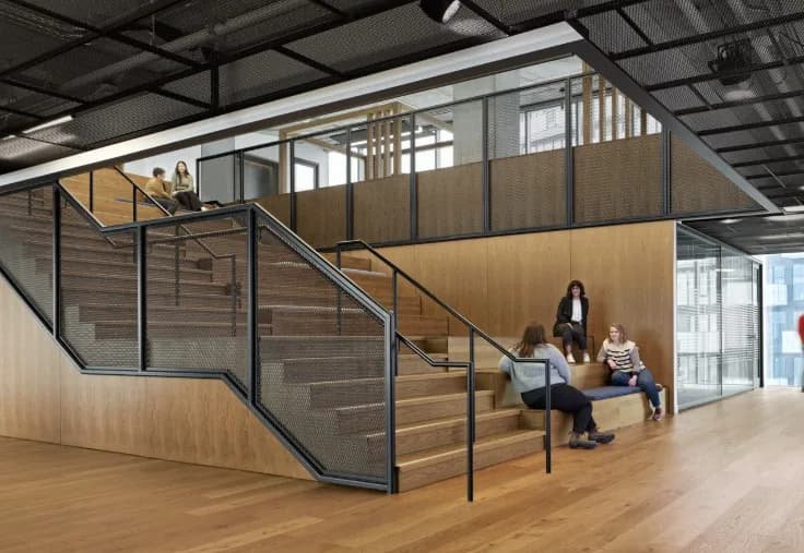 The Well, Toronto: Wide timber stadium stair with black steel mesh guardrails and handrails in an open office, people seated on the steps
