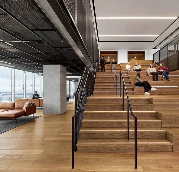 The Well, Toronto: Timber stadium stair with black steel handrails and a mesh guardrail, people seated on the upper steps, with a lounge area and windows beyond