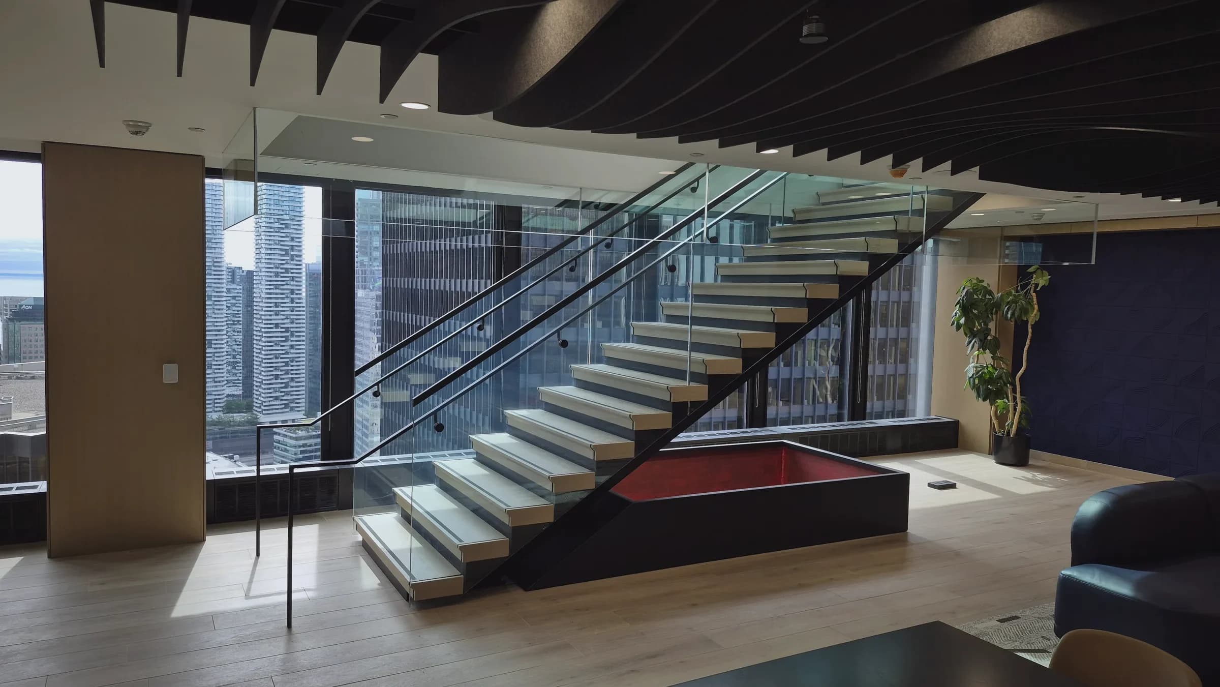 Office staircase, Toronto: Floating stair with light wood treads on a black steel stringer and glass guardrails, beside floor-to-ceiling windows over downtown towers