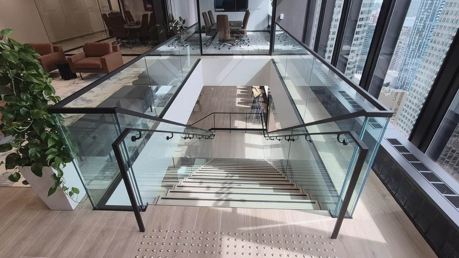 Office staircase, Toronto: View down a glass-guarded stair opening cut into an office floor, with black steel handrails and city towers through the windows